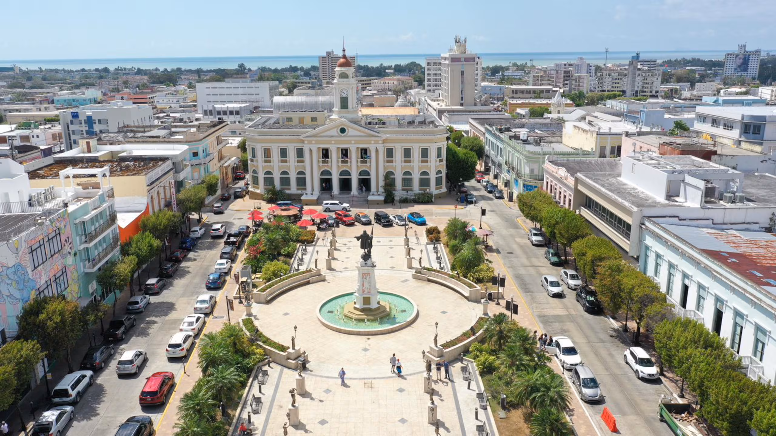 Aerial view of Plaza Colón, its central monument and fountain, and surrounding buildings in Mayagüez, with the coast in the distance.